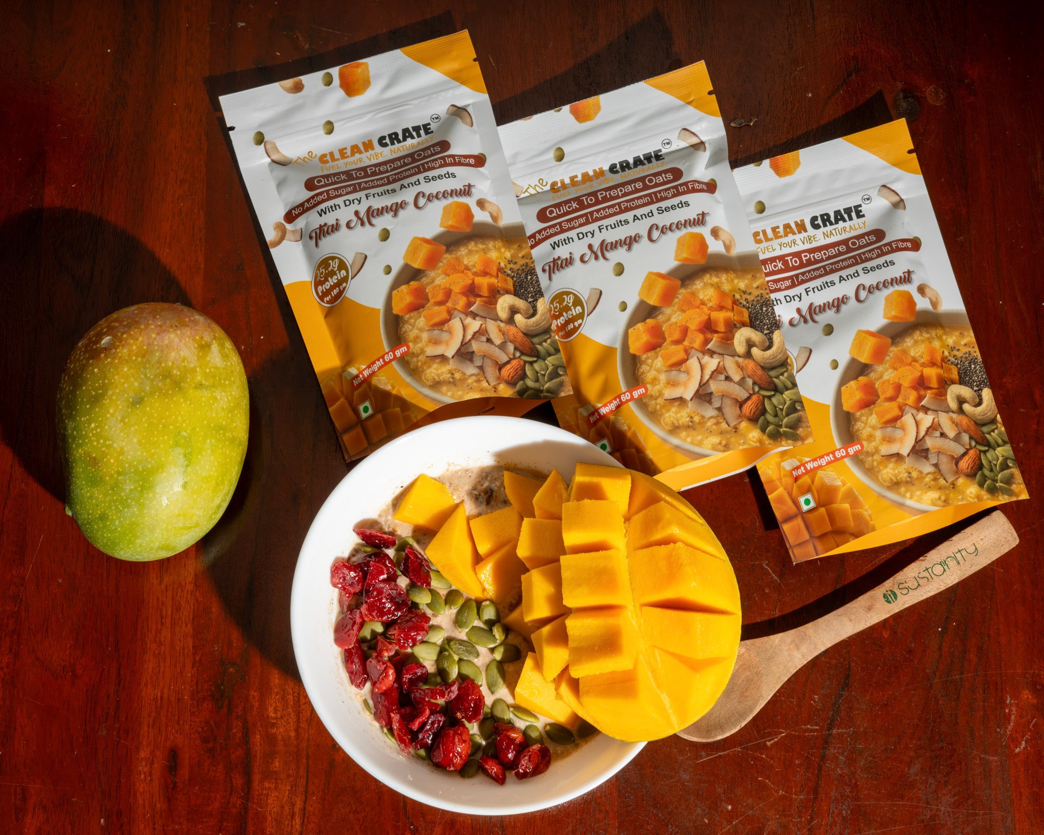 Three Thai Mango Coconut packs on a dark wooden table beside a whole mango and a bowl of prepared oats topped with mango and cranberries