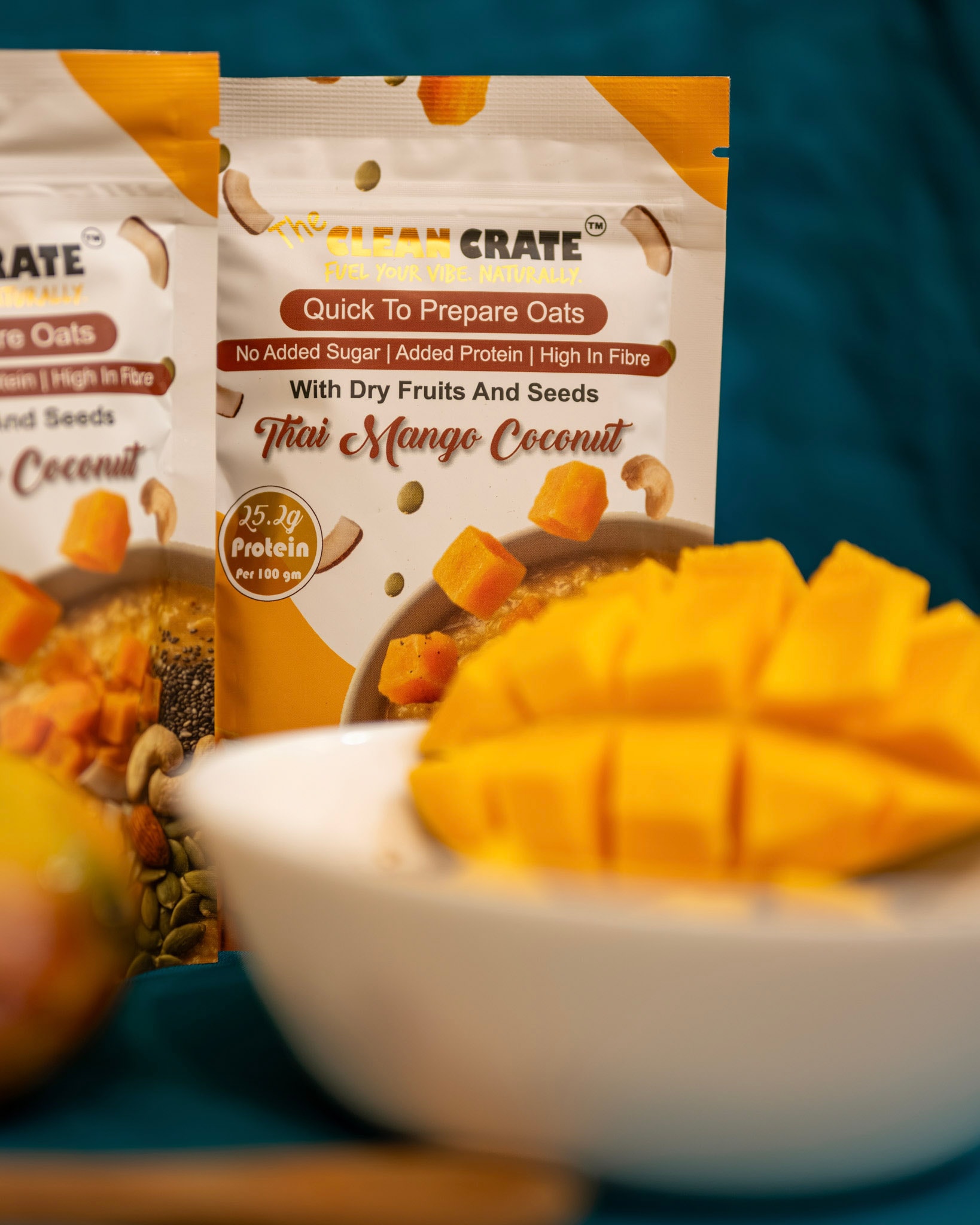 A Thai Mango Coconut pack photographed close up against a deep teal backdrop, with a bowl of cut mango in front of it